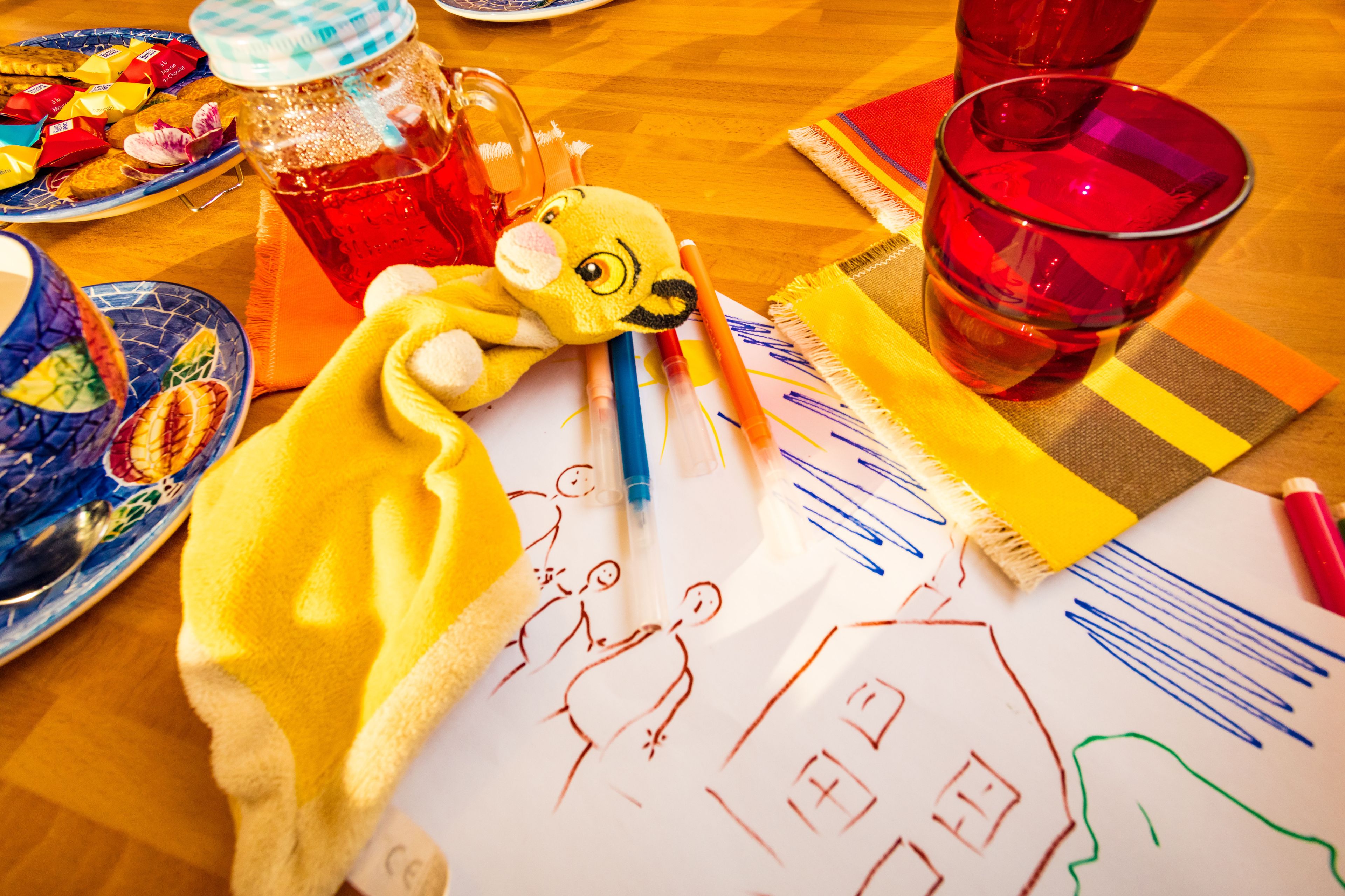 A table with a drawing, colored pencils, a plush toy, and a glass of red liquid.