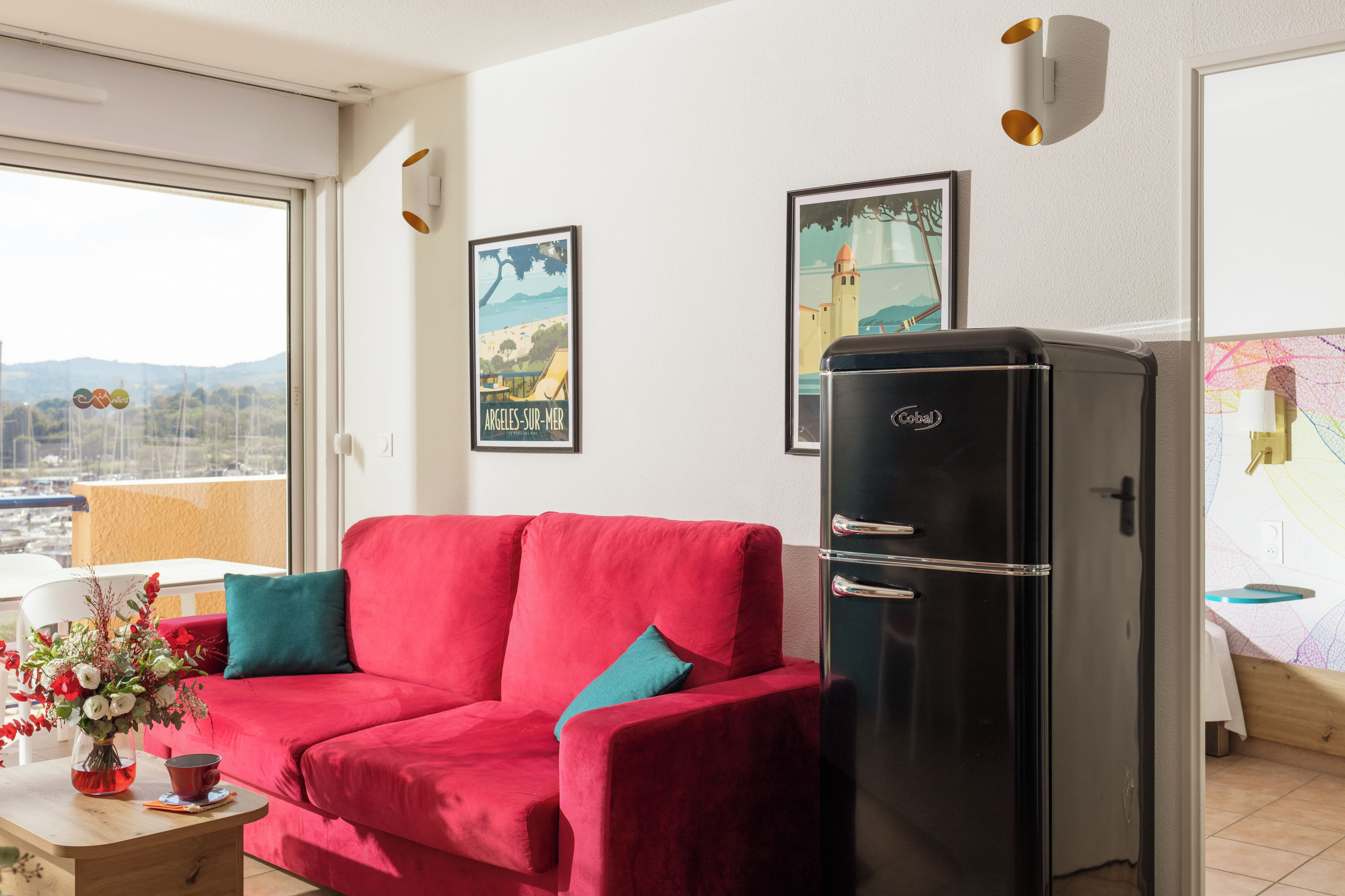 Un sofá rojo con cojines verde azulado, un refrigerador negro y una ventana con vistas a colinas.