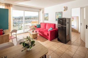 Una sala moderna con un sofá rojo, una mesa de comedor de madera, un refrigerador y vistas a un puerto deportivo a través de amplios ventanales.