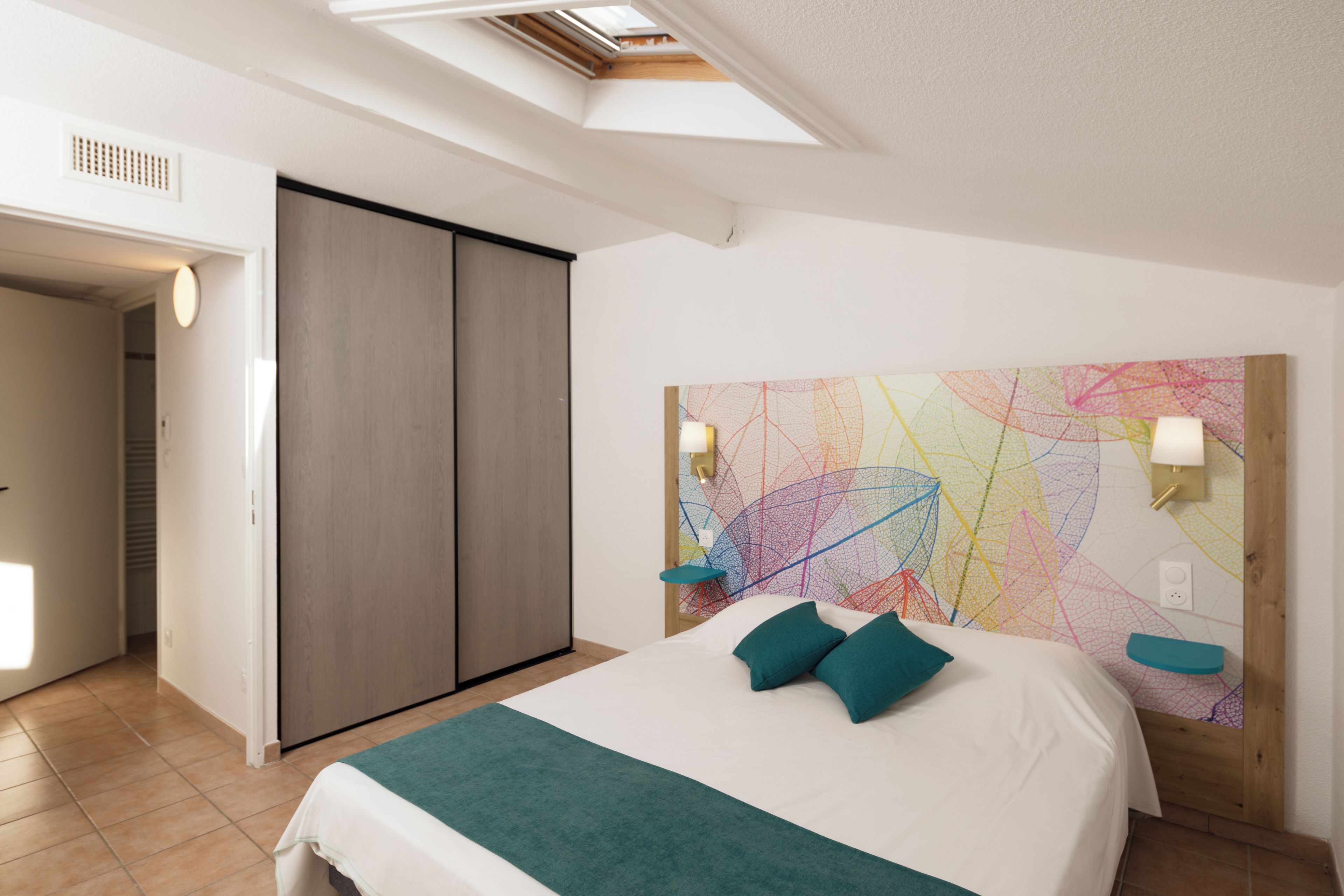 Un dormitorio moderno con tragaluz, una cama grande con almohadas turquesa, un colorido cuadro abstracto en la pared y un cabecero de madera.