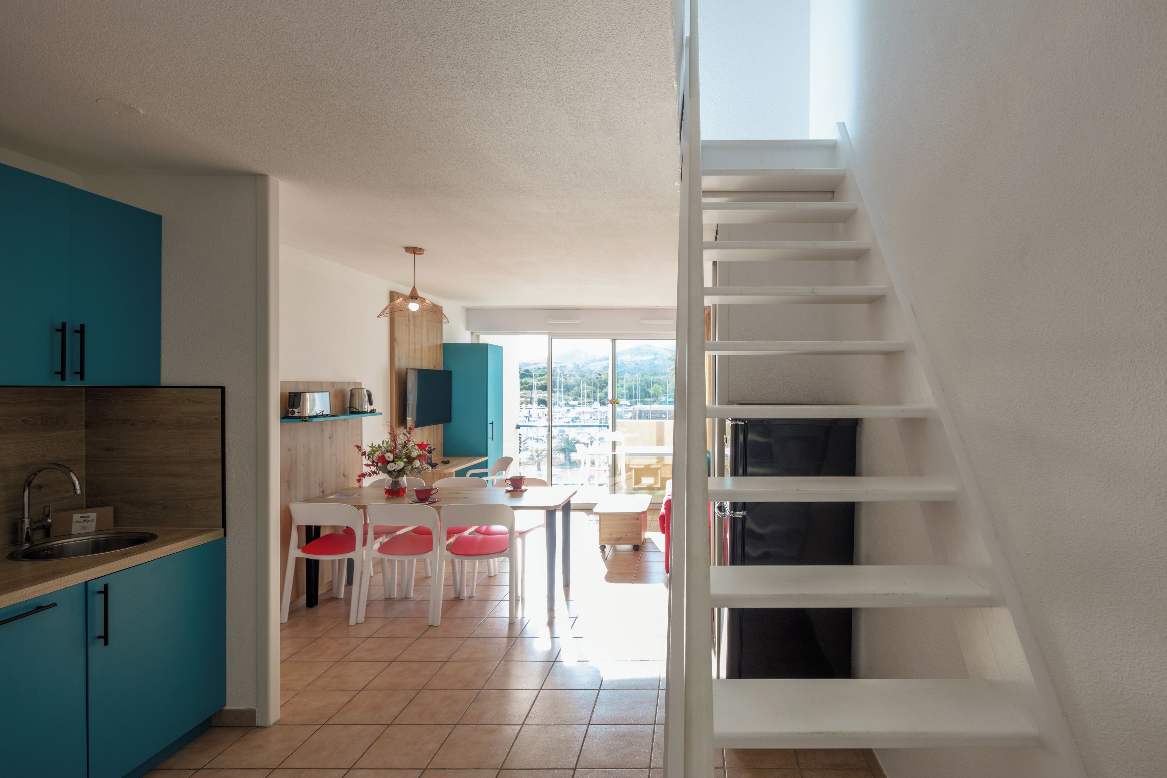 Una cocina moderna con armarios color turquesa, un comedor con sillas blancas y una escalera que conduce a un nivel superior.