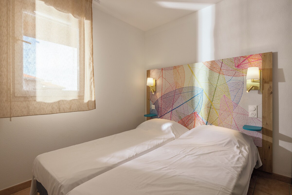 Una habitación con dos camas individuales, una ventana con cortina, un cuadro abstracto y colorido en la pared, y dos lámparas de pared.