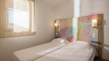 Una habitación con dos camas individuales, una ventana con cortina, un cuadro abstracto y colorido en la pared, y dos lámparas de pared.