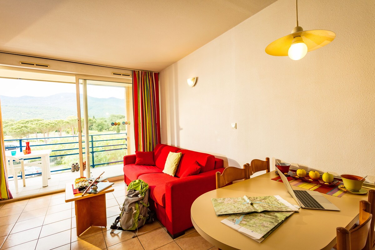 Una habitación con un sofá rojo, una mesa de comedor con una computadora portátil y frutas, y vistas a zonas verdes a través de amplios ventanales.