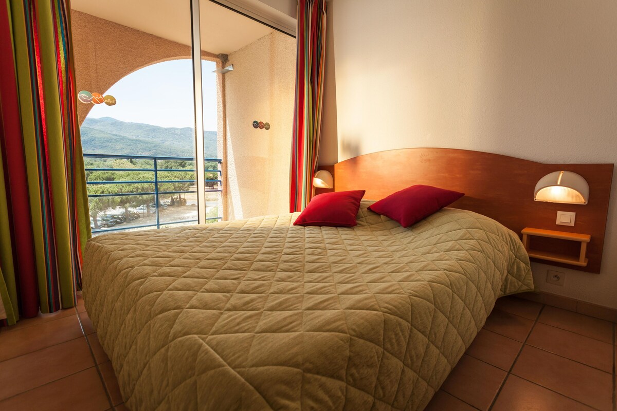 Una cama con cabecera acolchada, dos almohadas, una mesita de noche con lámpara y vistas a un balcón con barandilla.