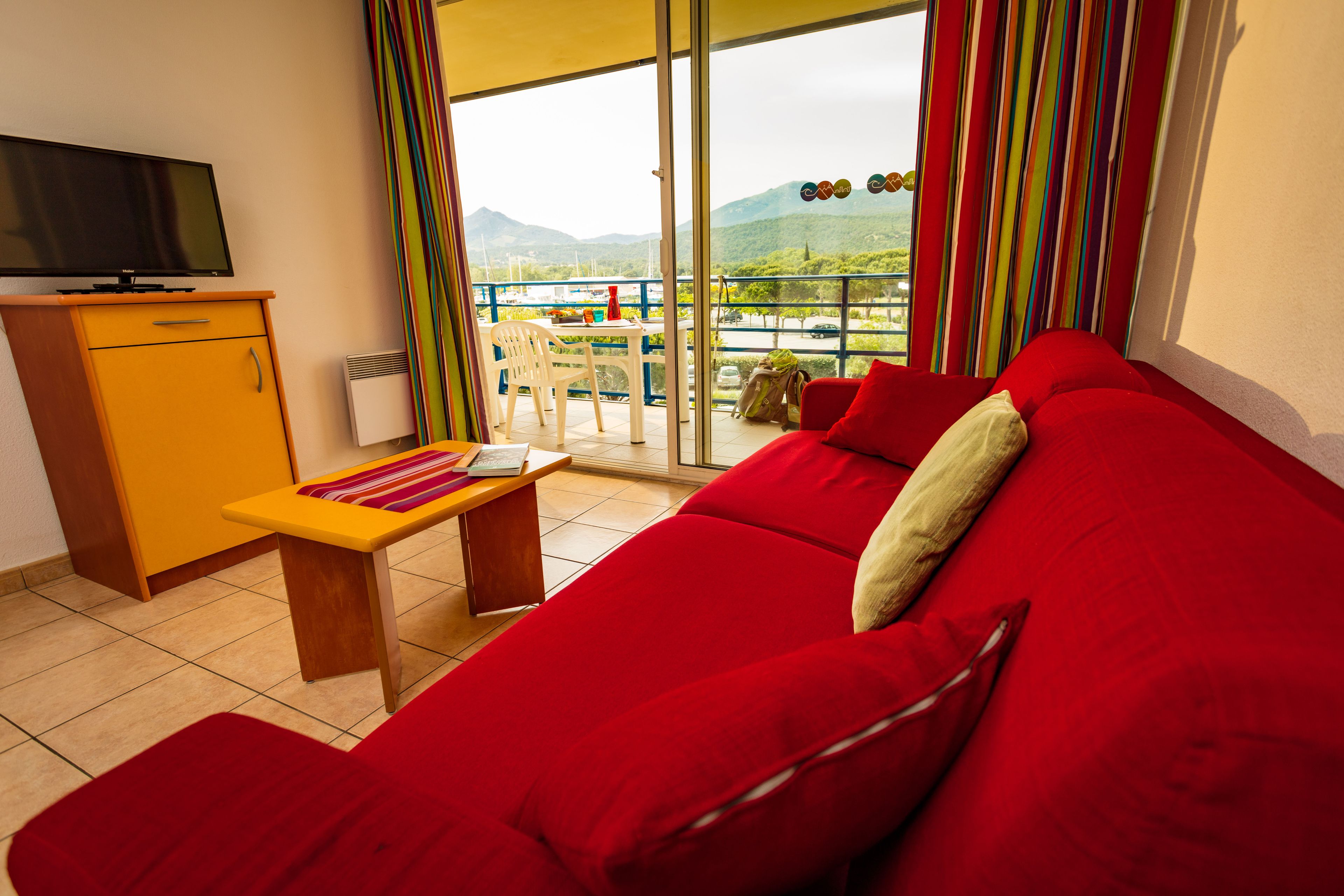 Un sofá rojo, una mesita de madera, un televisor sobre un mueble bajo y un balcón con vistas a montañas.