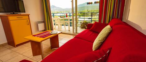 Un sofá rojo, una mesita de madera, un televisor sobre un mueble bajo y un balcón con vistas a montañas.