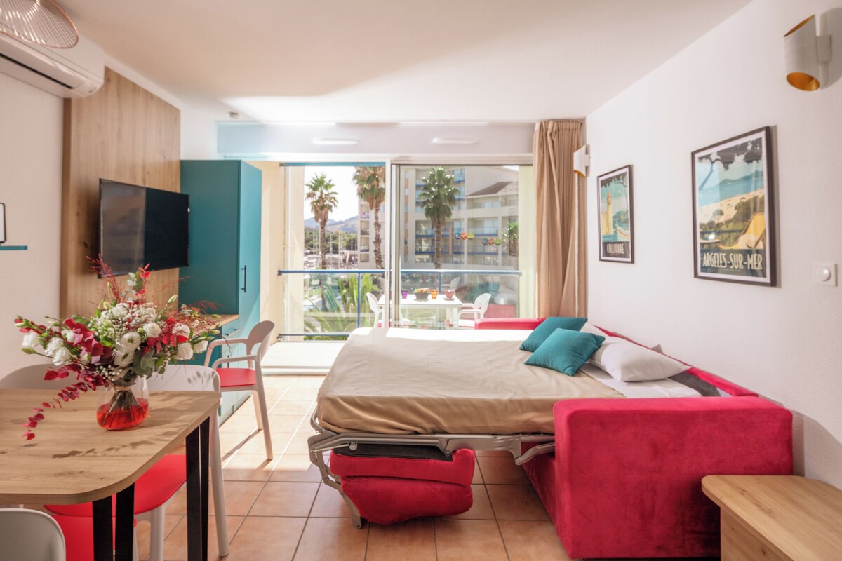 Habitación de hotel con cama, televisión, mesa de comedor con sillas y balcón con vistas a palmeras y edificios.