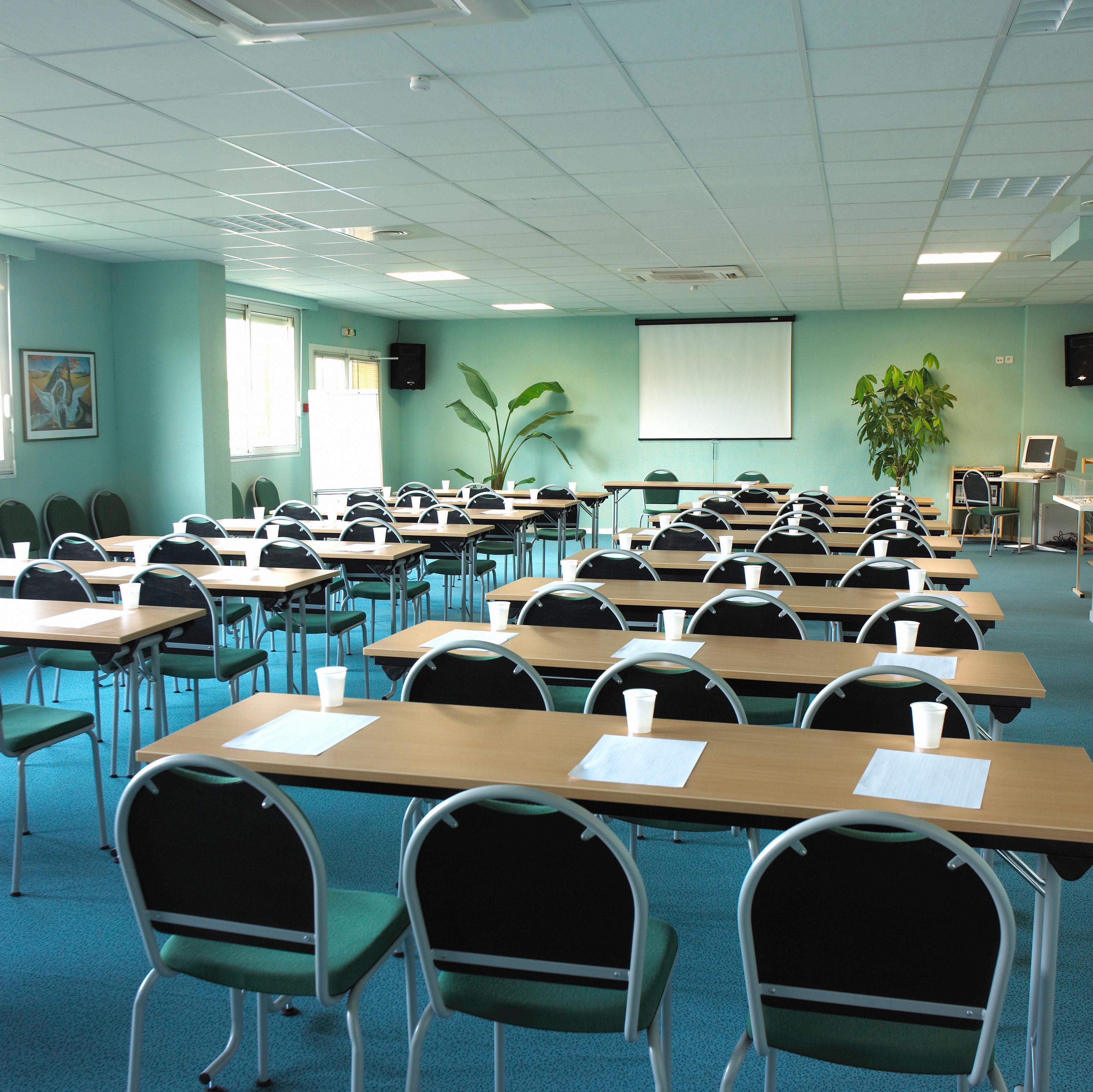 Un aula con filas de escritorios, sillas, una pantalla de proyector y una planta.