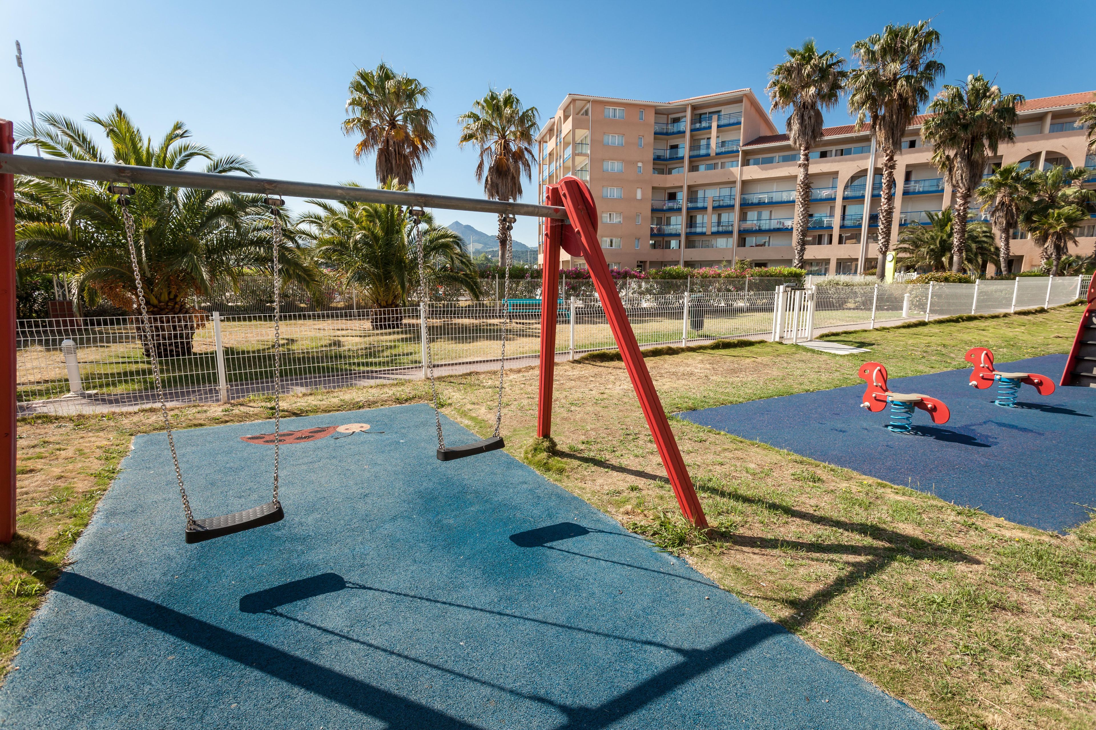 Un parque infantil con columpios y sube y bajas, rodeado por una cerca, palmeras y un edificio al fondo.