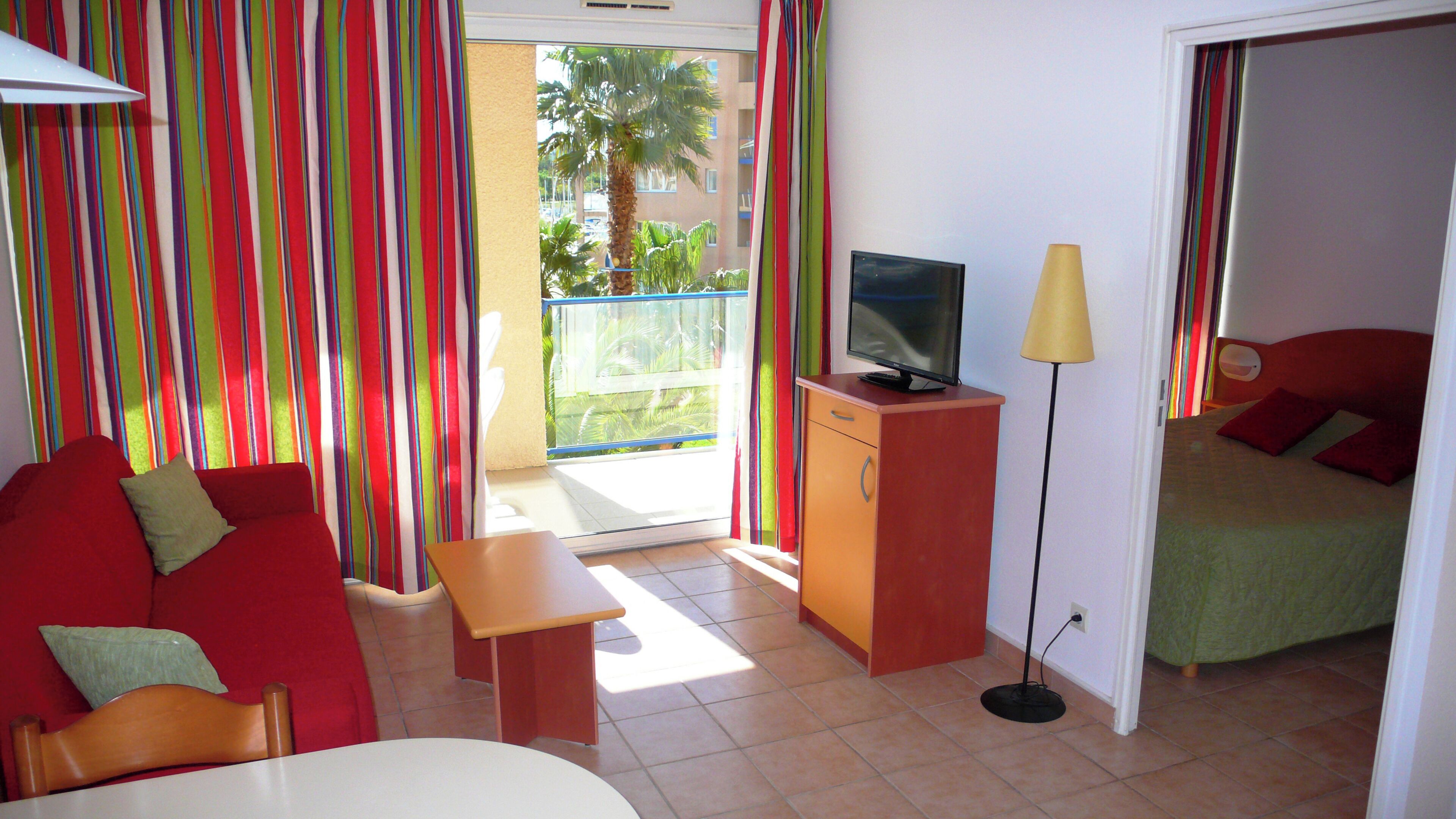 A hotel room with a red sofa, a small table, a TV on a wooden stand, a lamp, and a bed with red pillows.
