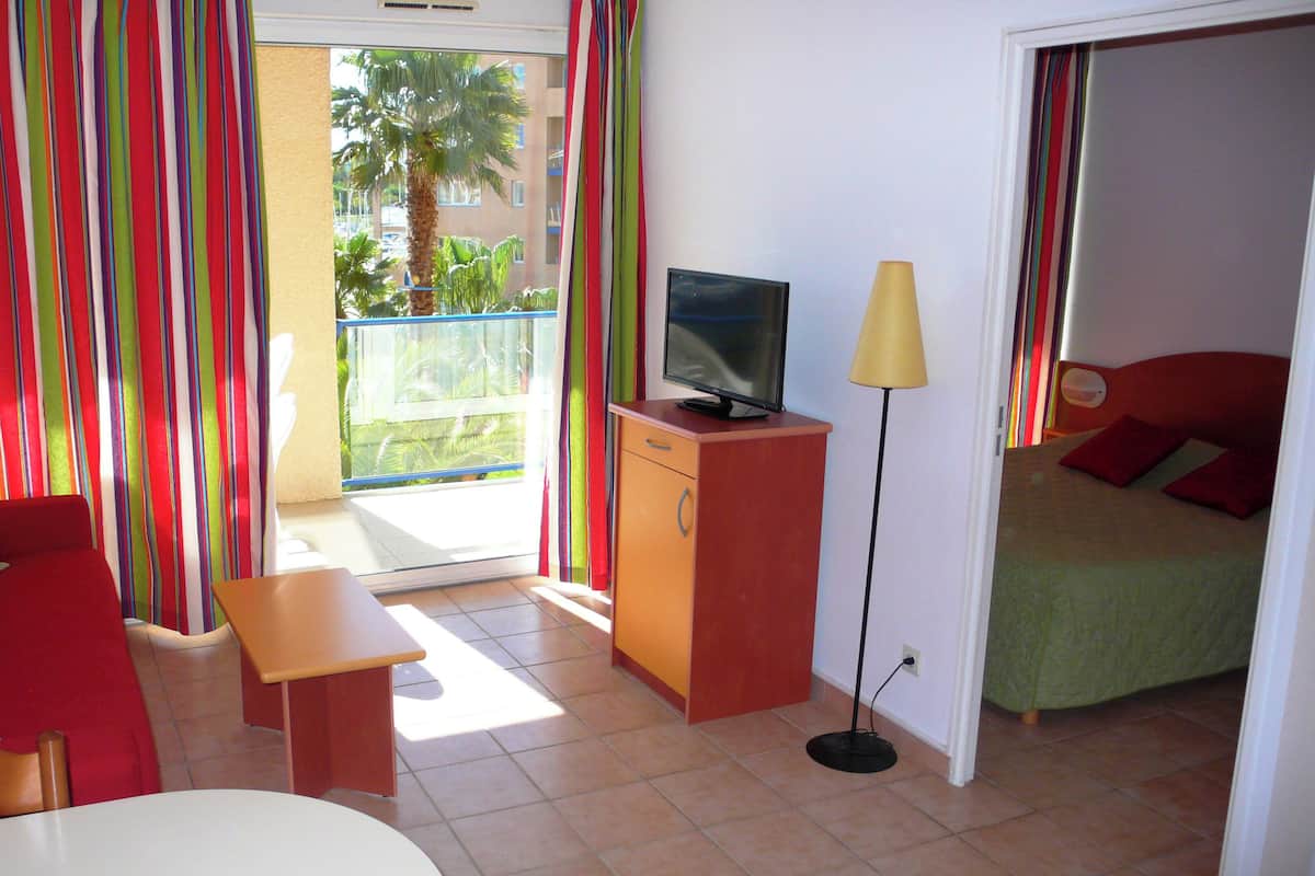 A hotel room with a red sofa, a small table, a TV on a wooden stand, a lamp, and a bed with red pillows.