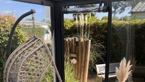 Una habitación con una silla colgante, una mesita con un florero y vistas a un jardín a través de amplios ventanales.