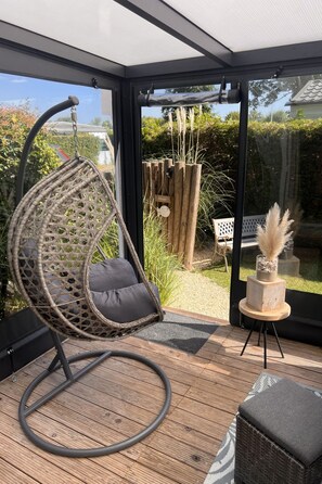 A room with a hanging chair, a small table with a vase, and a view of a garden through large windows. - RBR 852 - Beach Resort Kamperland by Interhome (Kamperland)