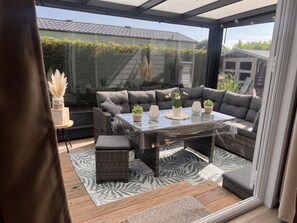 Een overdekt terras met een glazen tafel, grijze zitting met kussens en potplanten.