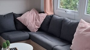 Una sala moderna con un sofá modular, una mesa de centro redonda de madera y una mesita auxiliar. Se ve una ventana con vistas a zonas verdes.