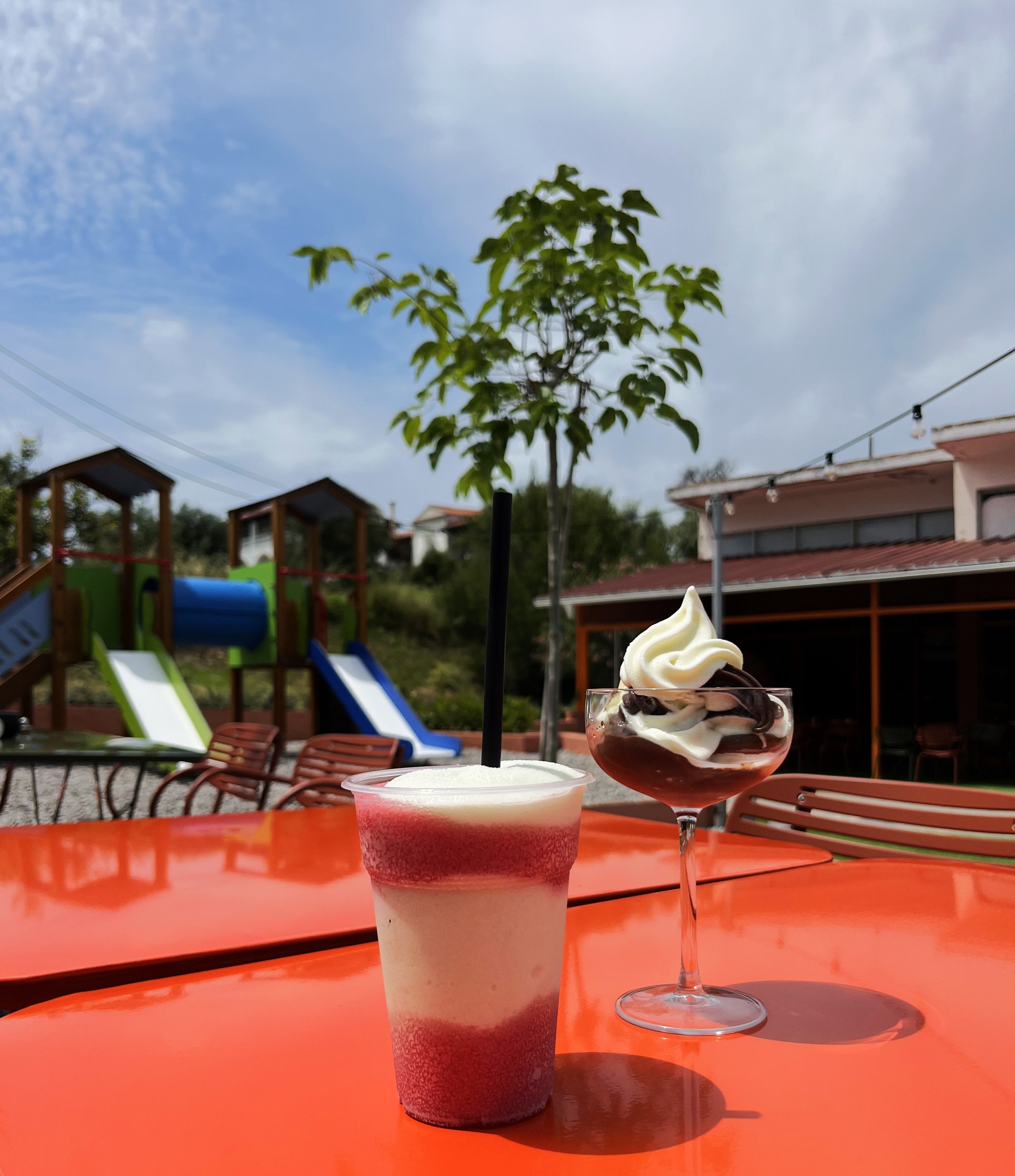 Una mesa con una bebida y un postre, un parque infantil y un edificio al fondo.