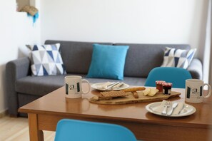 Un comedor con una mesa de madera, platos, cubiertos y una taza. Un sofá gris con cojines estampados se ve al fondo.