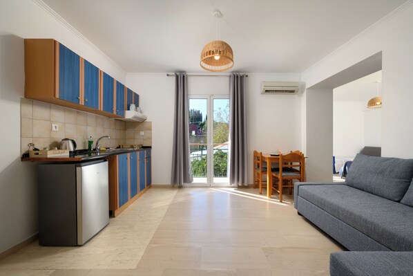 A modern kitchen with a built-in island, a dining area with a wooden table and chairs, and a grey sofa. - Aquarius Beach Hotel (Corfu)