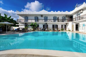 Una piscina con sillas de descanso y sombrillas, un edificio blanco con balcones y un cielo azul despejado.
