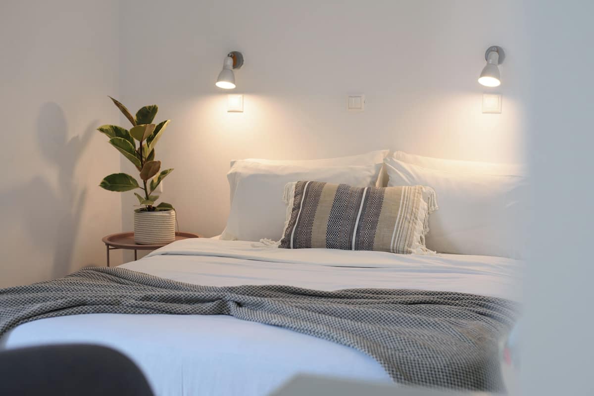 A neatly made bed with a striped pillow, a potted plant, and two wall-mounted lights.