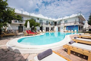 A swimming pool with lounge chairs and umbrellas, a building with balconies, and a sign reading 'Corfu Greece.'