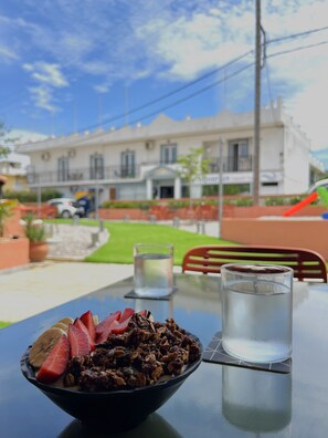 A table with a bowl of food, two glasses of water, and a view of a building with a car and greenery. - Aquarius Beach Hotel (Corfu)