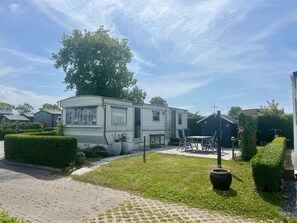 Ein Mobilheim mit einer Terrasse, einem großen Baum und einem gepflegten Rasen.