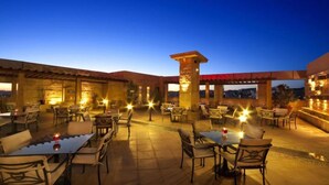 An outdoor dining area with tables and chairs, a stone pillar, and a view of the cityscape at dusk.