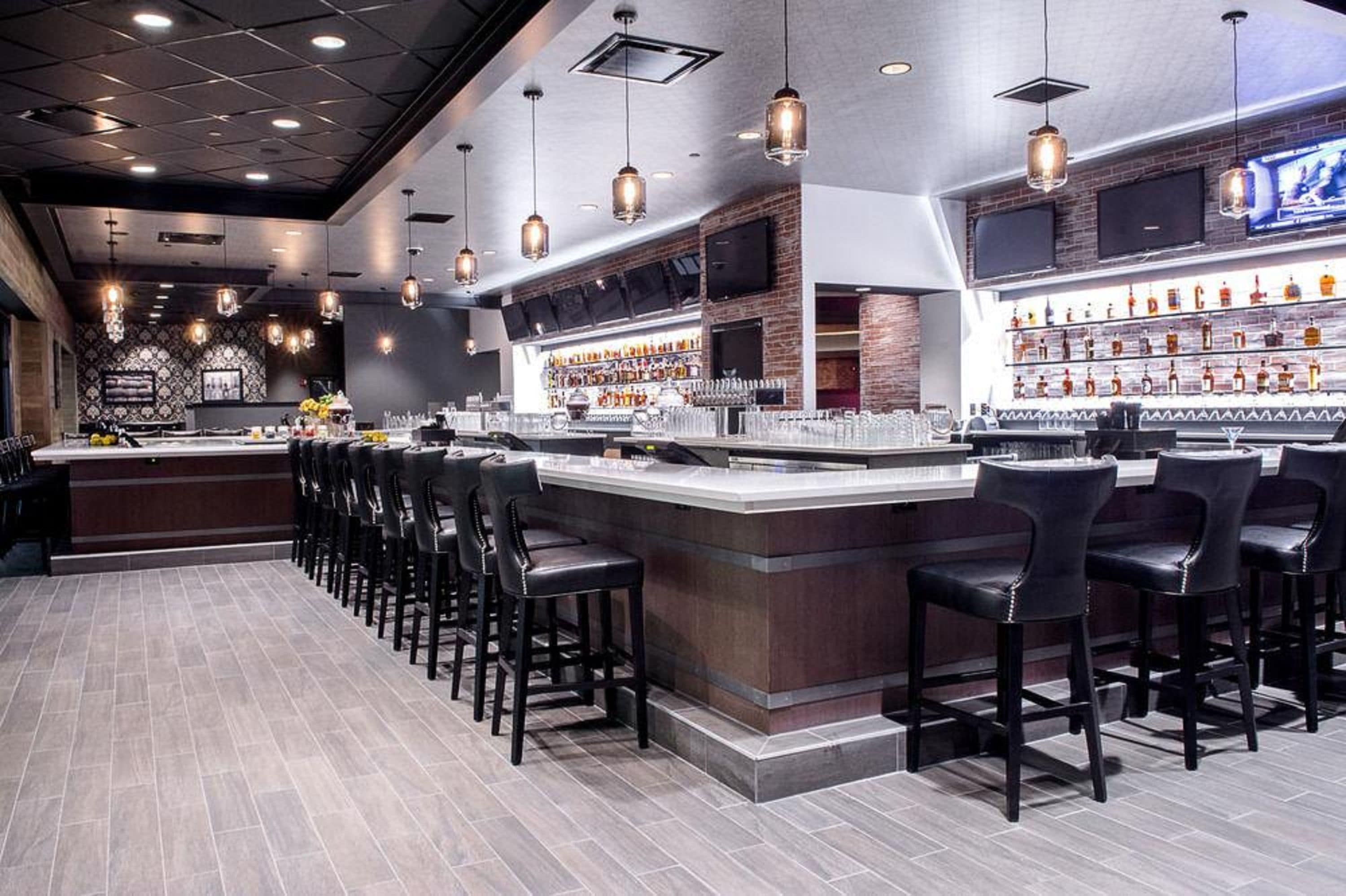 A bar area with a long counter, multiple bar stools, and a well-stocked shelf with bottles.