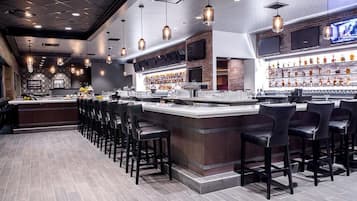 A bar area with a long counter, multiple bar stools, and a well-stocked shelf with bottles.