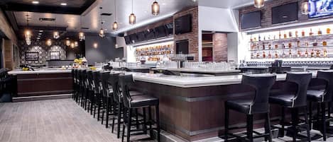 A bar area with a long counter, multiple bar stools, and a well-stocked shelf with bottles.