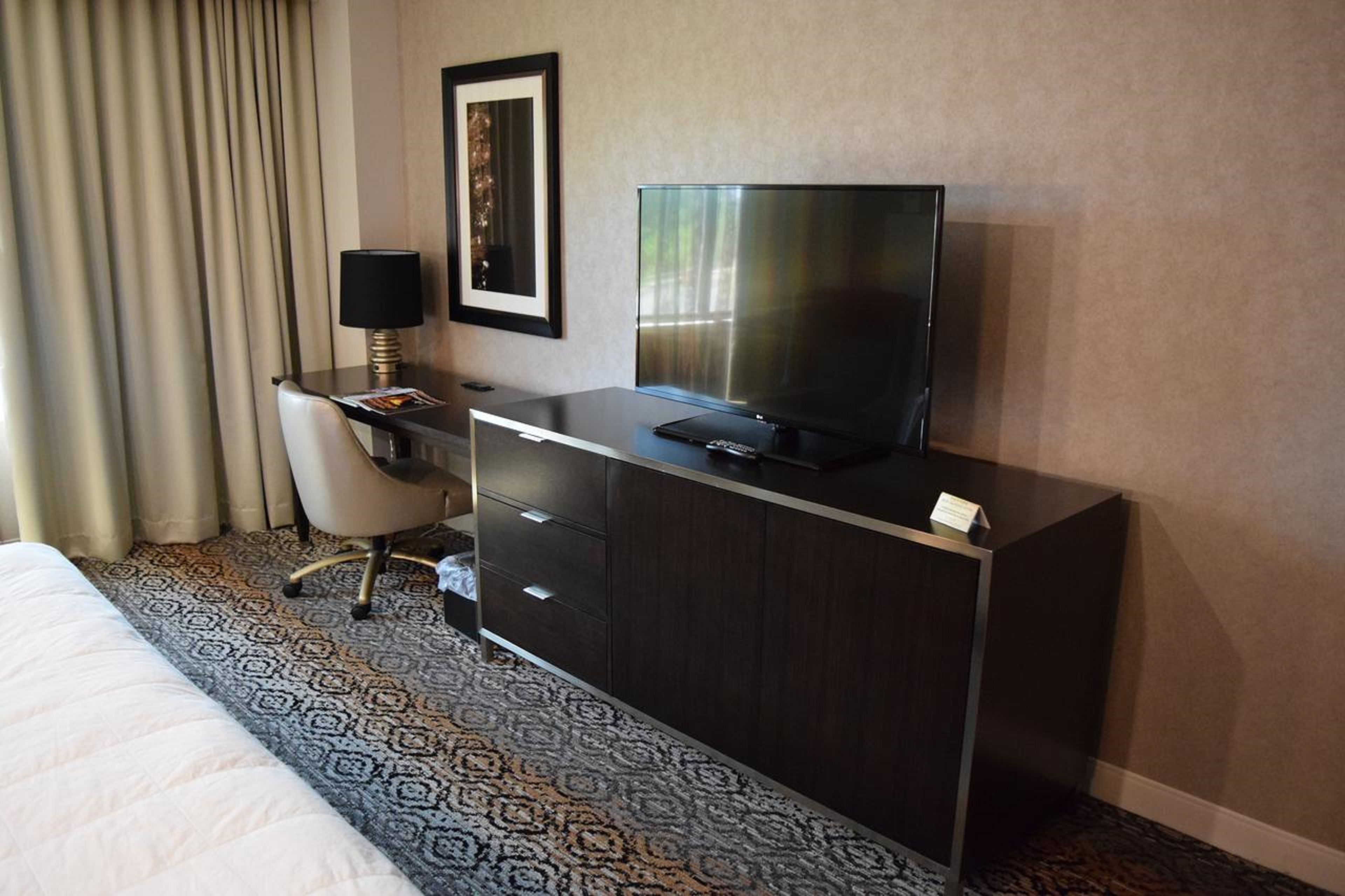 A hotel room with a bed, a desk with a TV, a chair, and a framed picture on the wall.