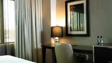 A hotel room with a bed, a desk with a lamp, a chair, a framed picture on the wall, and a window with curtains.