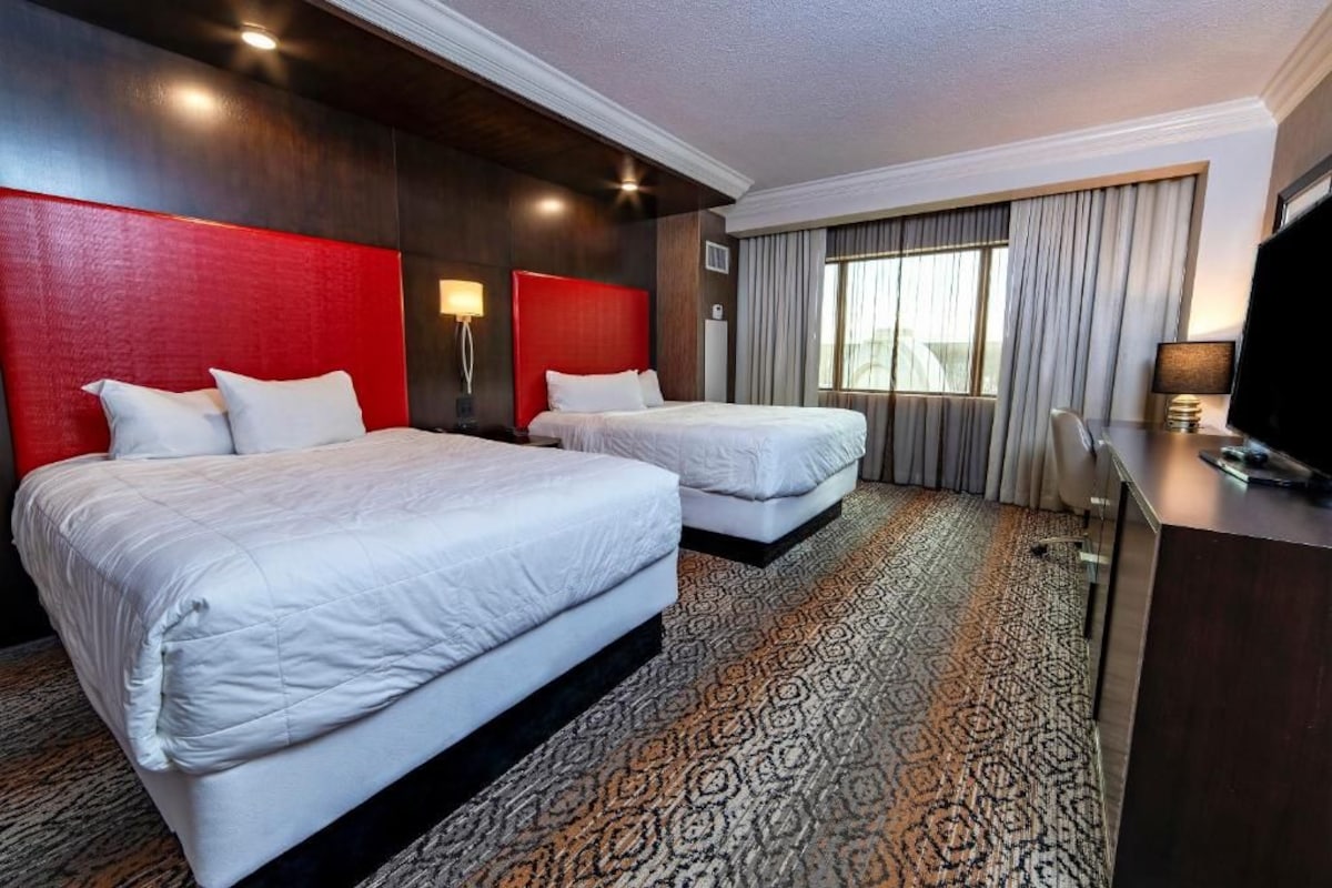 A hotel room with two beds, a desk, a TV, and a large window with curtains.