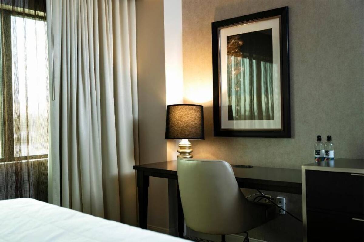 A hotel room with a bed, a desk with a lamp, a chair, a framed picture on the wall, and a window with curtains.