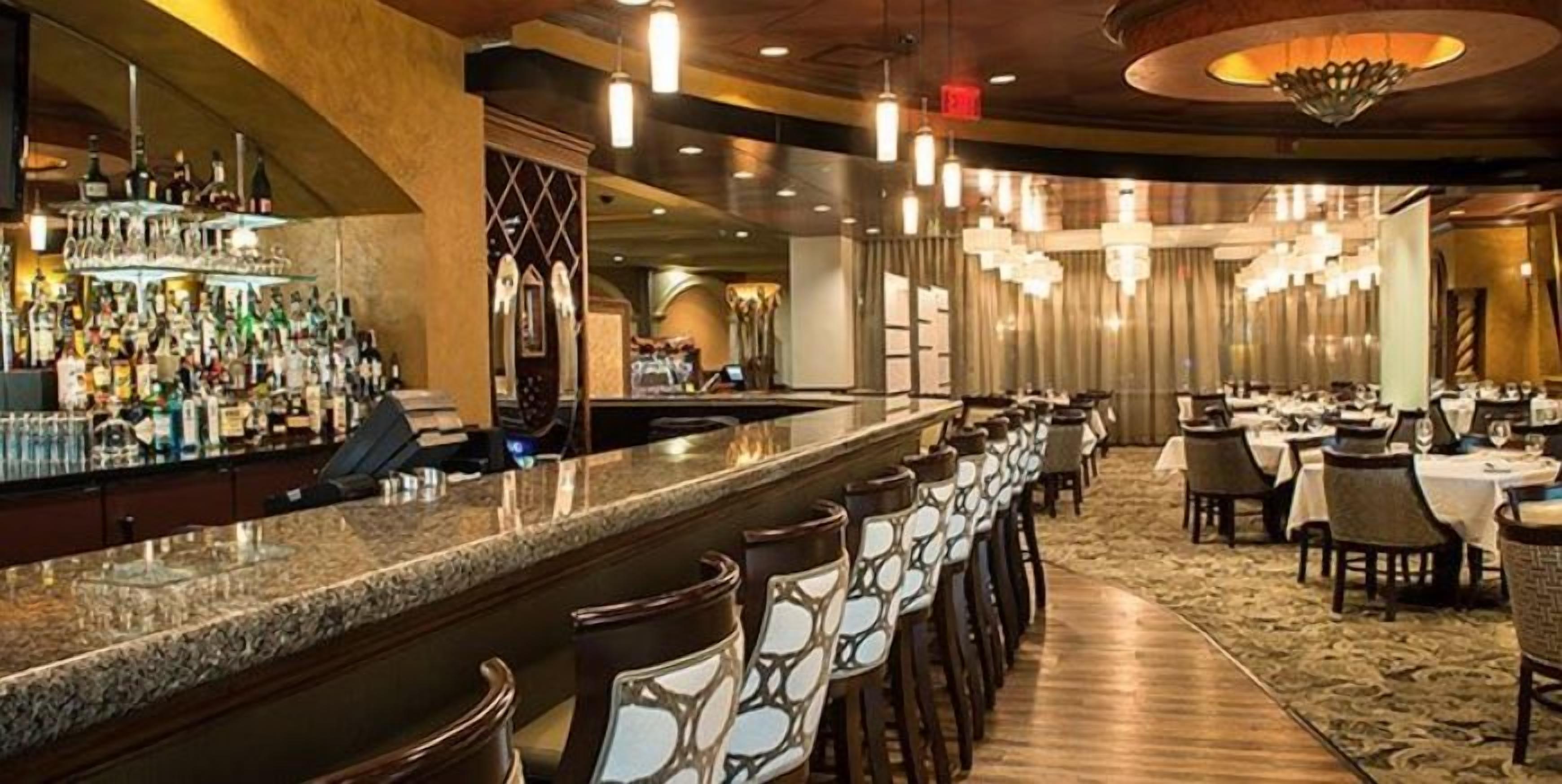 A bar area with a long counter and multiple chairs, a dining area with tables and chairs, and a chandelier.