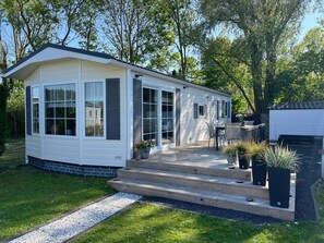 Ein eingeschossiges Mobilheim mit überdachter Terrasse, einer kleinen Sitzgelegenheit im Freien und einem gepflasterten Zufahrtsweg.