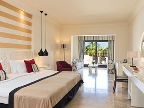 A hotel room with a large bed, a desk, a chair, a sofa, and a balcony with a view of palm trees. - Juweira Boutique Hotel (Salalah)