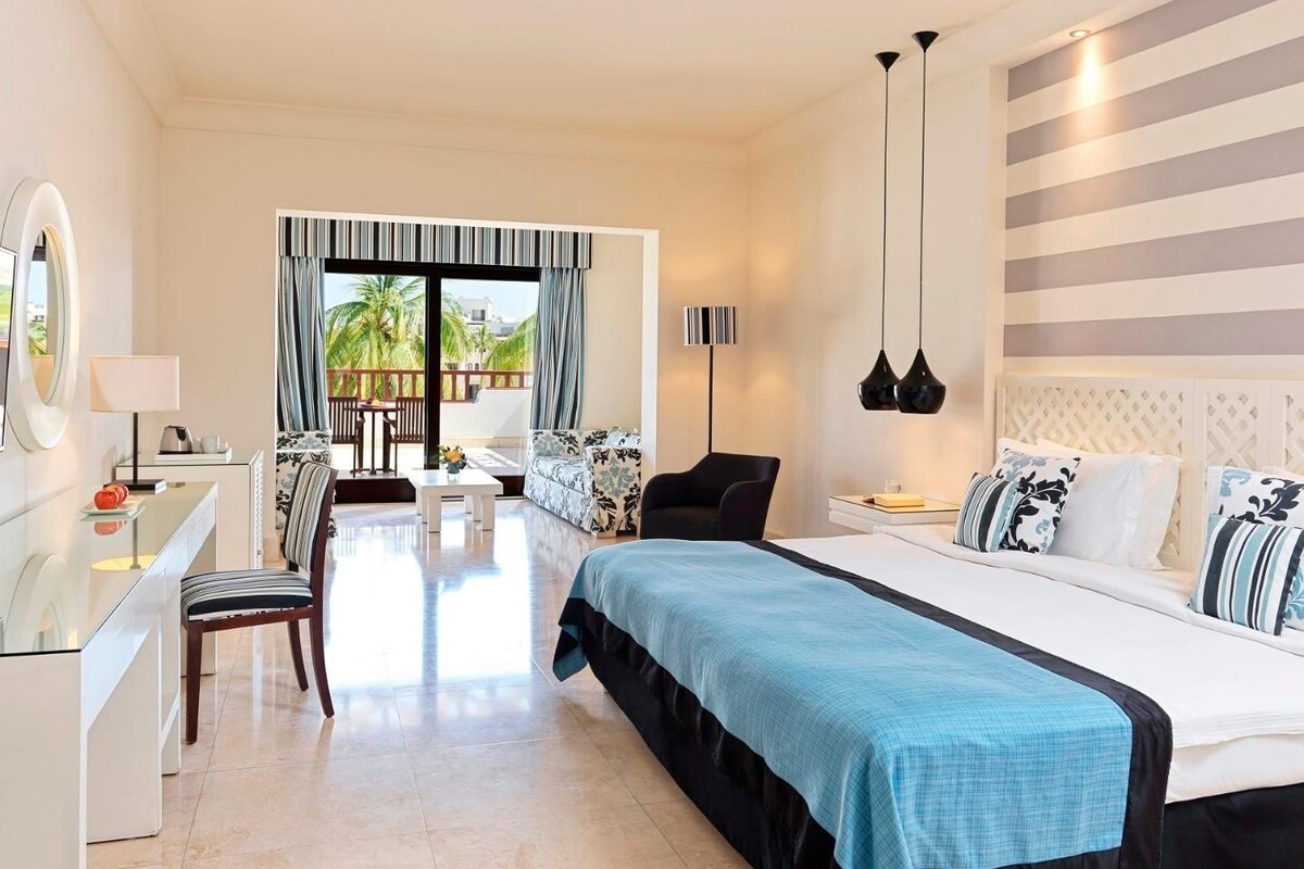 A hotel room with a bed, a desk, a chair, a sofa, and a view of palm trees.