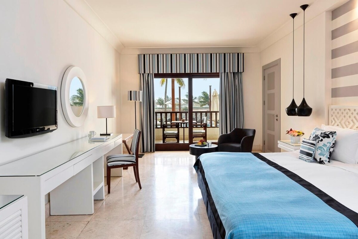 A hotel room with a bed, a desk with a TV, a chair, a small table with a lamp, and a balcony with a view.