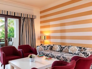 A hotel room with a sofa, two armchairs, a coffee table with a vase, a lamp, and a view of palm trees through the window. - Juweira Boutique Hotel (Salalah)