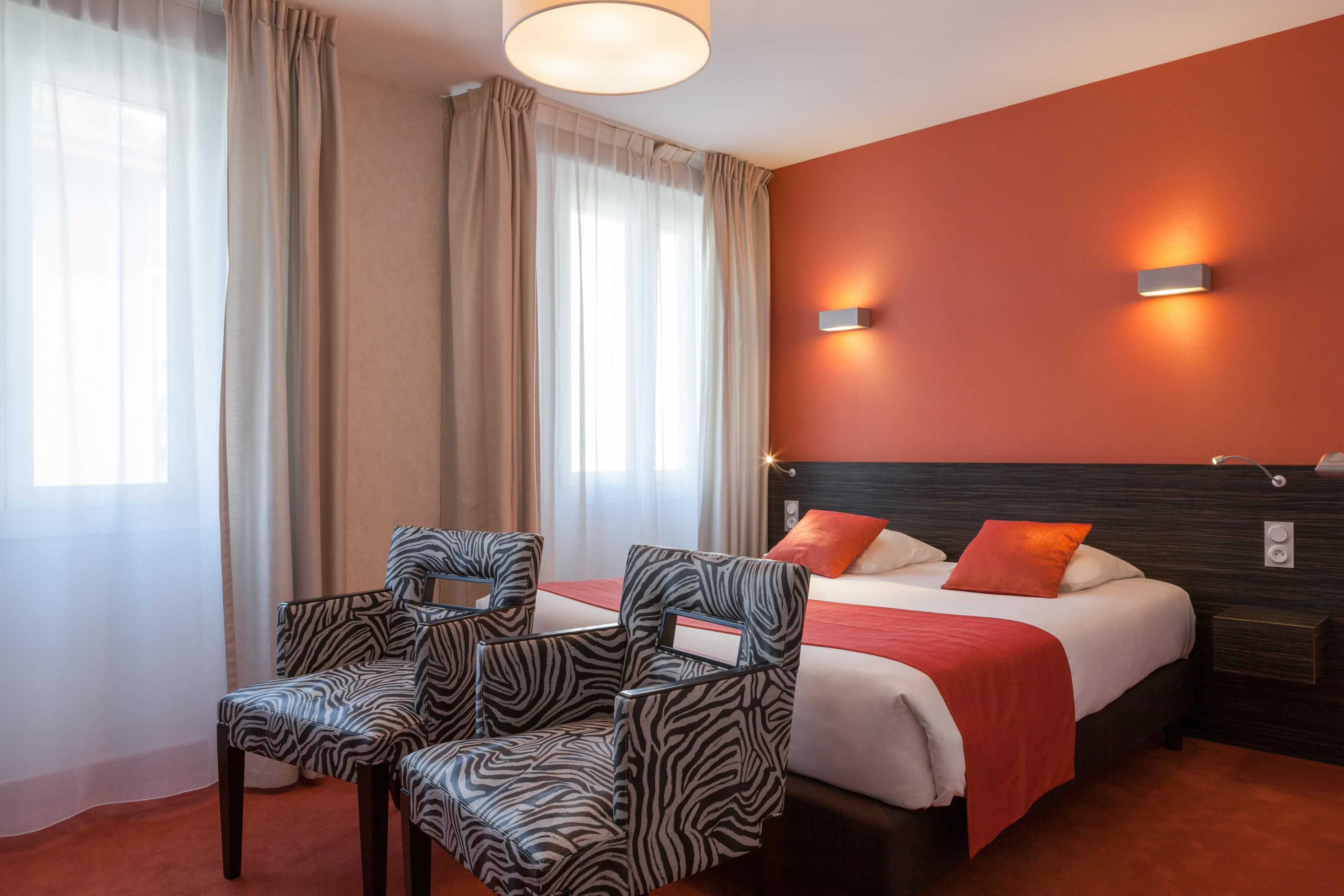 Habitación de hotel con cama, dos sillas, una mesita de noche y una lámpara en la pared.