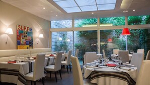 Un restaurante con mesas dispuestas para comer, un mural con temática de 'SURF' y grandes ventanales que ofrecen vistas a zonas verdes.