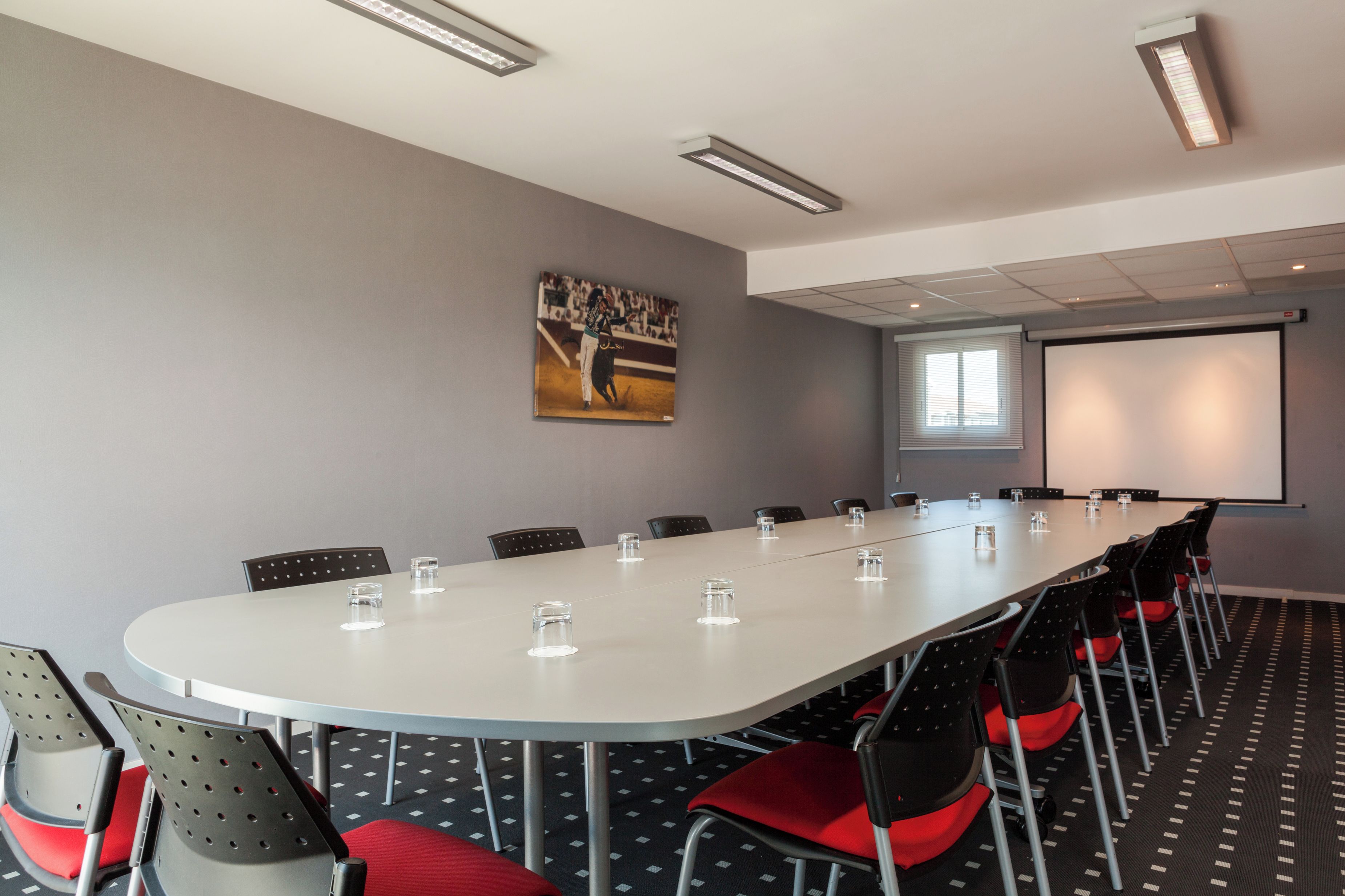 Un salón de conferencias con una larga mesa, sillas, una pantalla de proyector y un cuadro en la pared.