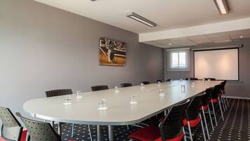 Un salón de conferencias con una larga mesa, sillas, una pantalla de proyector y un cuadro en la pared.