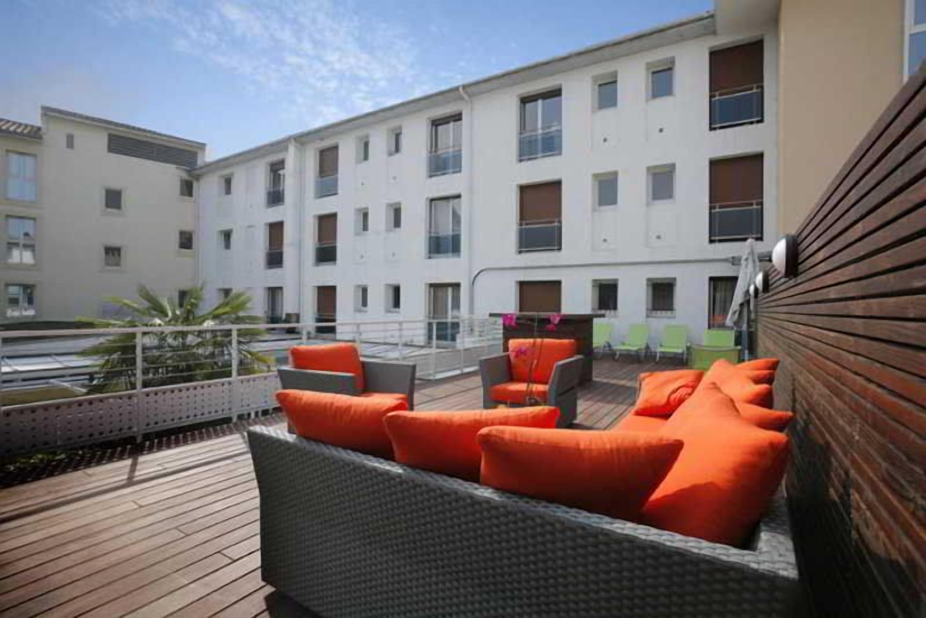 Une terrasse sur le toit, aménagée avec des meubles en osier et des coussins orange, d’où l’on peut admirer un immeuble doté de nombreuses fenêtres.