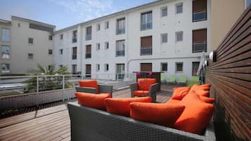 Une terrasse sur le toit, aménagée avec des meubles en osier et des coussins orange, d’où l’on peut admirer un immeuble doté de nombreuses fenêtres.