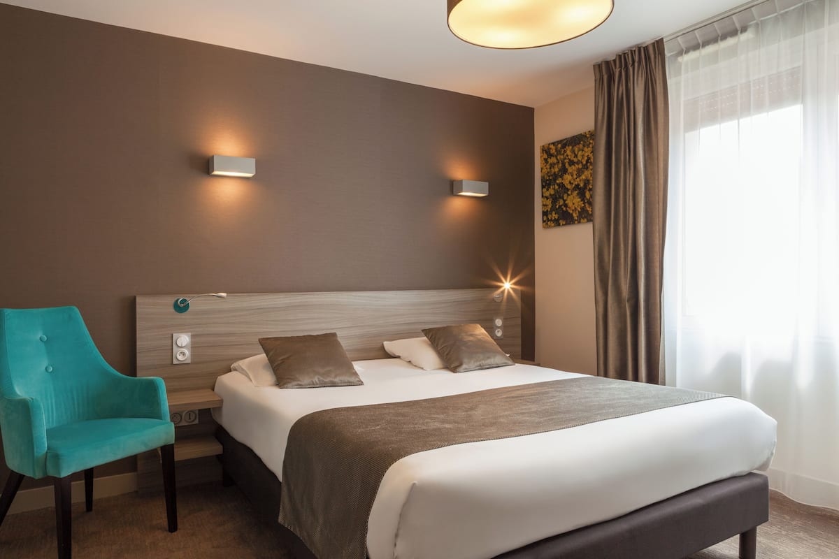 Une chambre d’hôtel avec un lit, un fauteuil de couleur turquoise, deux lampes fixées au mur et une fenêtre avec des rideaux.
