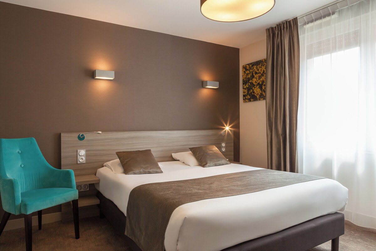 Une chambre d’hôtel avec un lit, un fauteuil de couleur turquoise, deux lampes fixées au mur et une fenêtre avec des rideaux.